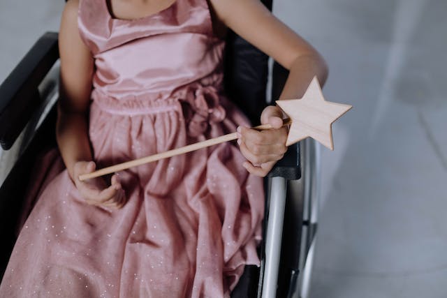 Girl wearing a pink dress, holding a wand, and sitting in a wheelchair